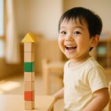 積み木を積み上げて振り返りながら嬉しそうに笑う幼い子どもの様子を、自然光の室内で撮影した写真。