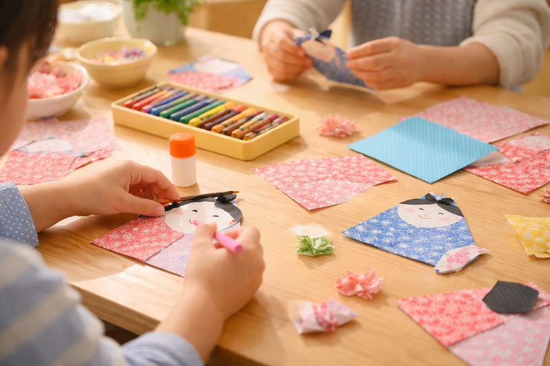 保育園のテーブルでひな祭り制作をしている日本人の園児たちの手元。折り紙や千代紙、クレヨン、のりが広がり、おひなさまの顔を描いている様子。