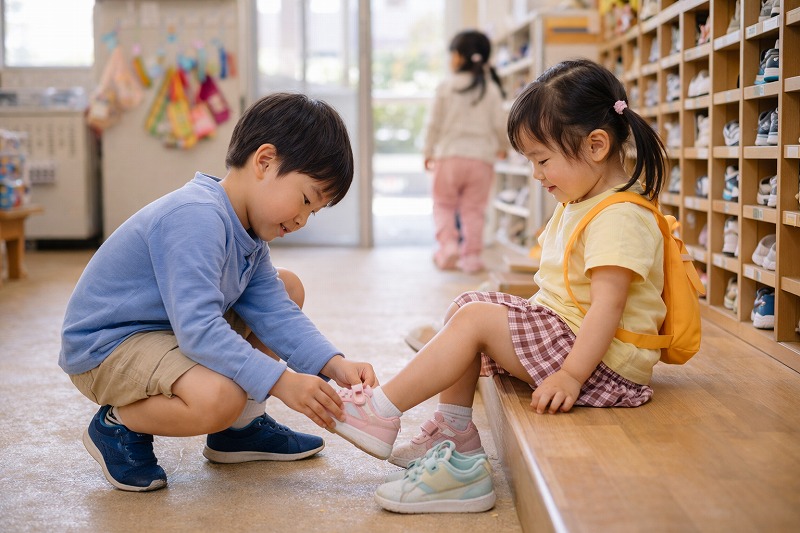 保育園の玄関で年長児が年下の子どもに靴を履かせてあげる優しい場面。思いやりや成長が感じられる、卒園前の子どもたちの姿。