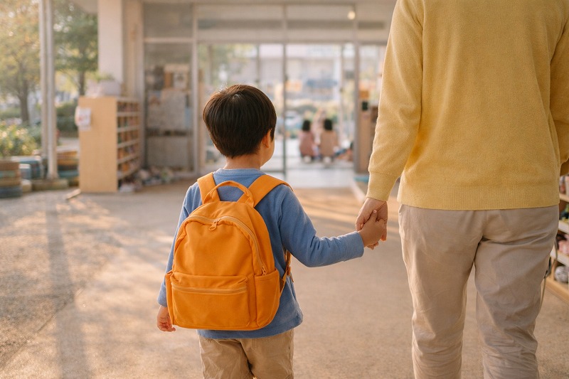 朝の保育園の玄関で親子が手をつないで歩く後ろ姿。登園時の温かな時間と、子どもの成長を見守る家族の想いが伝わるシーン。