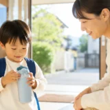 保育園の玄関で、小さな水筒のふたを自分で確認する幼児と、そばでやさしく見守る保護者