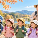 秋晴れの公園で、リュックを背負った保育園児たちが保育士と一緒に遠足を楽しんでいる様子。