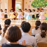 保育園のホールで、子どもたちが歌やダンスの発表をしており、後方から保護者が温かく見守る場面