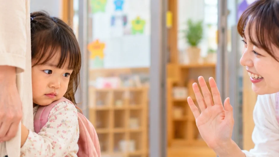 保育園の朝、先生に挨拶できず母親の後ろに隠れる幼児